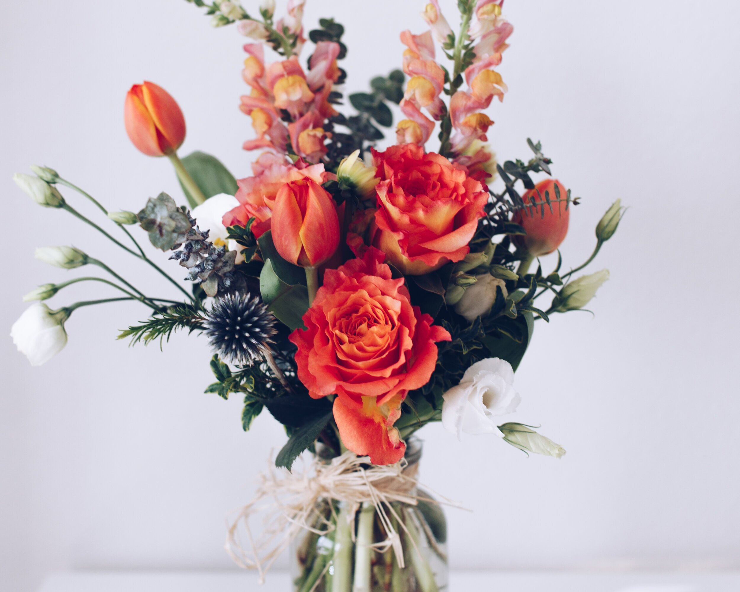 image-asset (7) Vibrant floral arrangement featuring orange roses, tulips, and snapdragons, complemented by greenery and white blooms, elegantly displayed in a glass vase with a natural twine accent. Perfect for home decor or gifting.